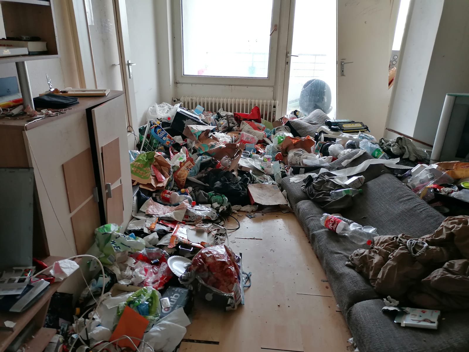 Unordentliches Zimmer mit Müll, Kleidung und Gegenständen auf dem Boden, Sofa und offenen Türen im Hintergrund.