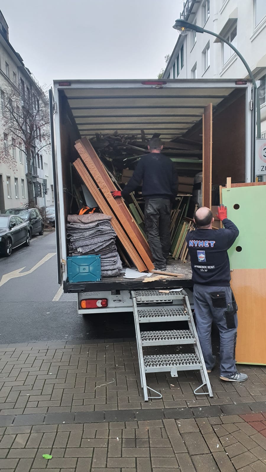 Zwei Männer verladen Holzbretter und Baumaterial in einem LKW, der am Straßenrand einer Stadt steht.