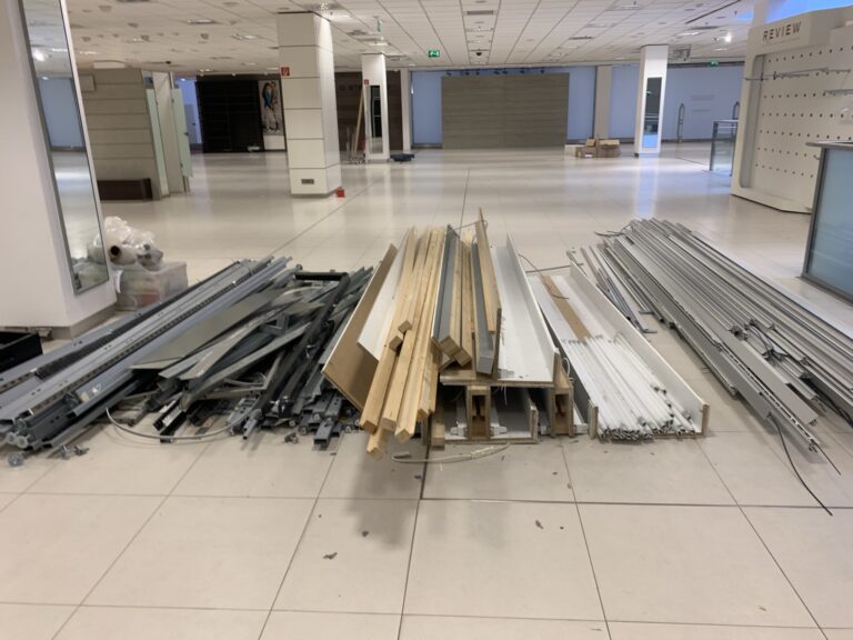 Metall- und Holzbalken, Platten und Rahmen sind ordentlich auf dem Boden in einer großen, leeren Gewerbefläche gestapelt.