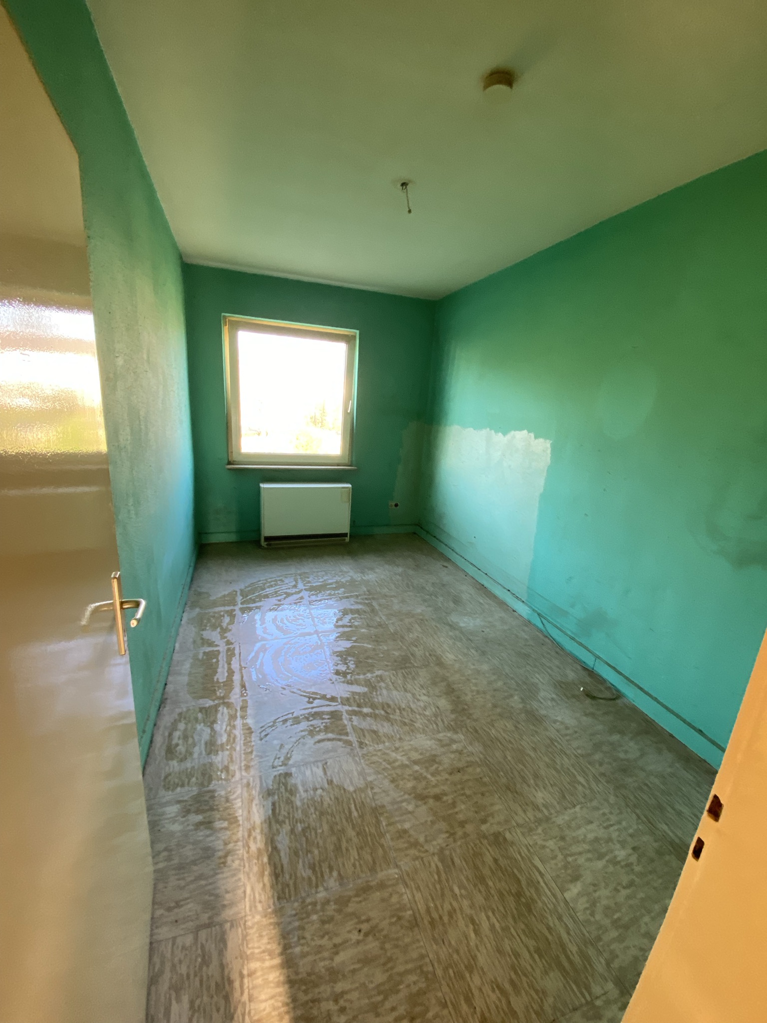 Teilweise überfluteter leerer Raum mit türkisfarbenen Wänden, Fliesenboden, Fenster, Heizkörper und Deckenleuchte.